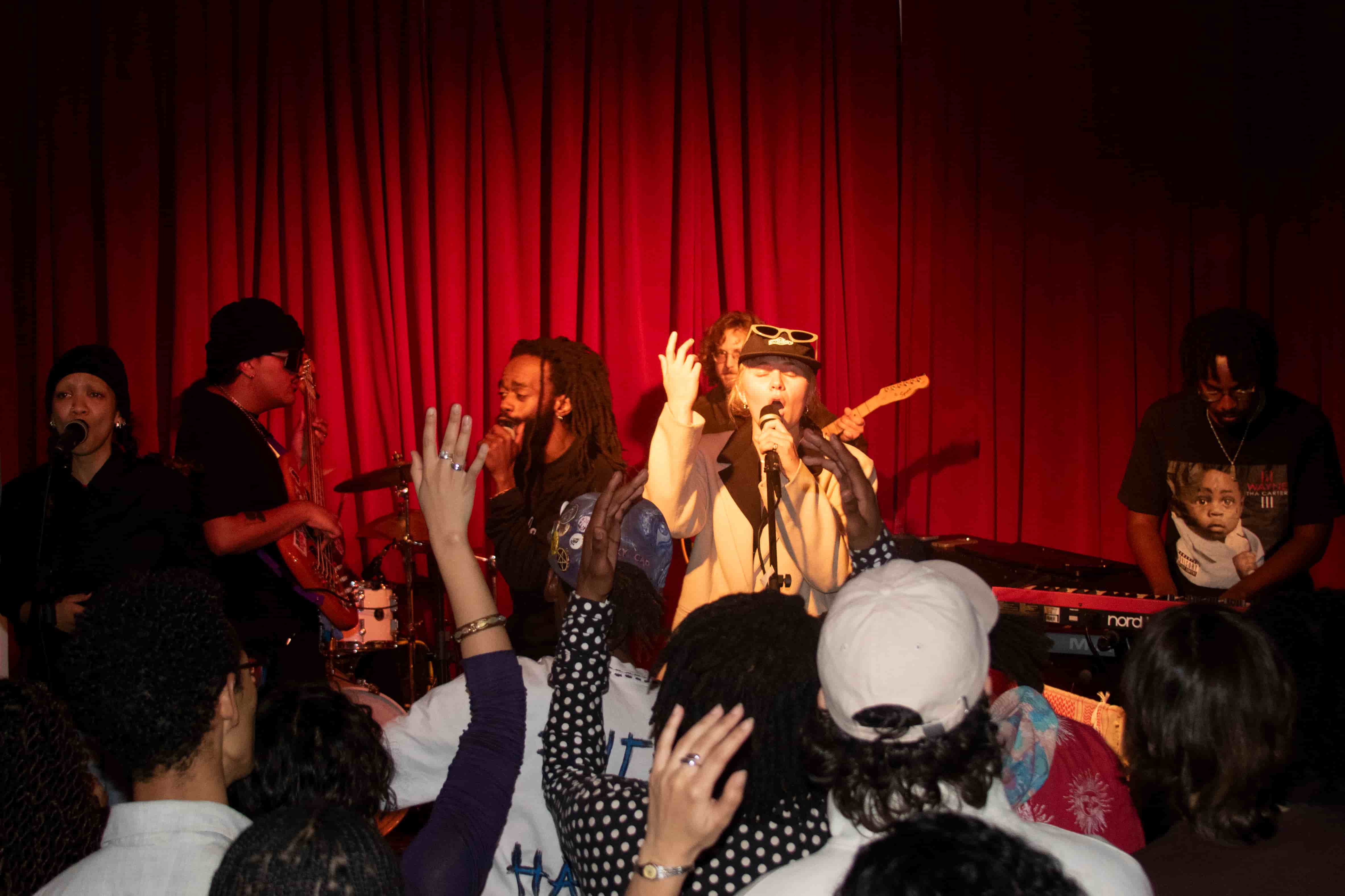 Full band on stage in front of a red curtain with crowd